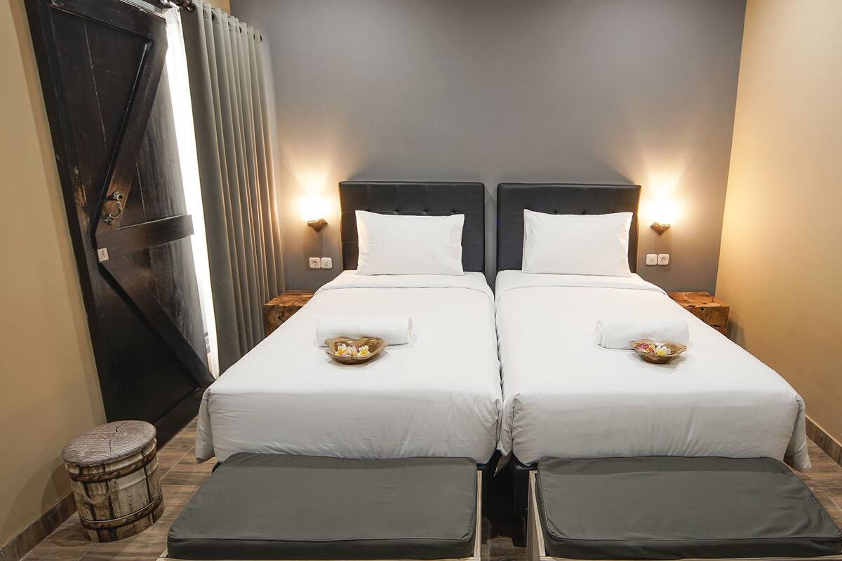 Habitación de hotel con dos camas, cada una con ropa blanca y almohadas, y mesitas de noche con lámparas.