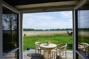 A conservatory with a view of a lake, a wooden deck, and a table with chairs. - Cottage Remise for 6 persons with large terrace - Gut Grubnow Rügen (Neuenkirchen)