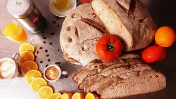 A loaf of bread, sliced bread, tomatoes, oranges, and two cups of coffee on a metallic surface.