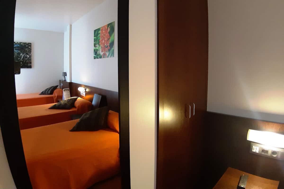 A hotel room with two orange beds, a wooden wardrobe, a painting on the wall, and a bedside table with a lamp.