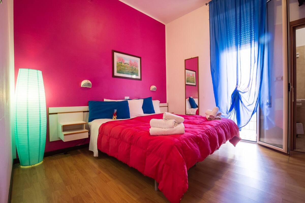 A hotel room with a red bedspread, a wooden floor, a large window with blue curtains, and a painting on the wall.