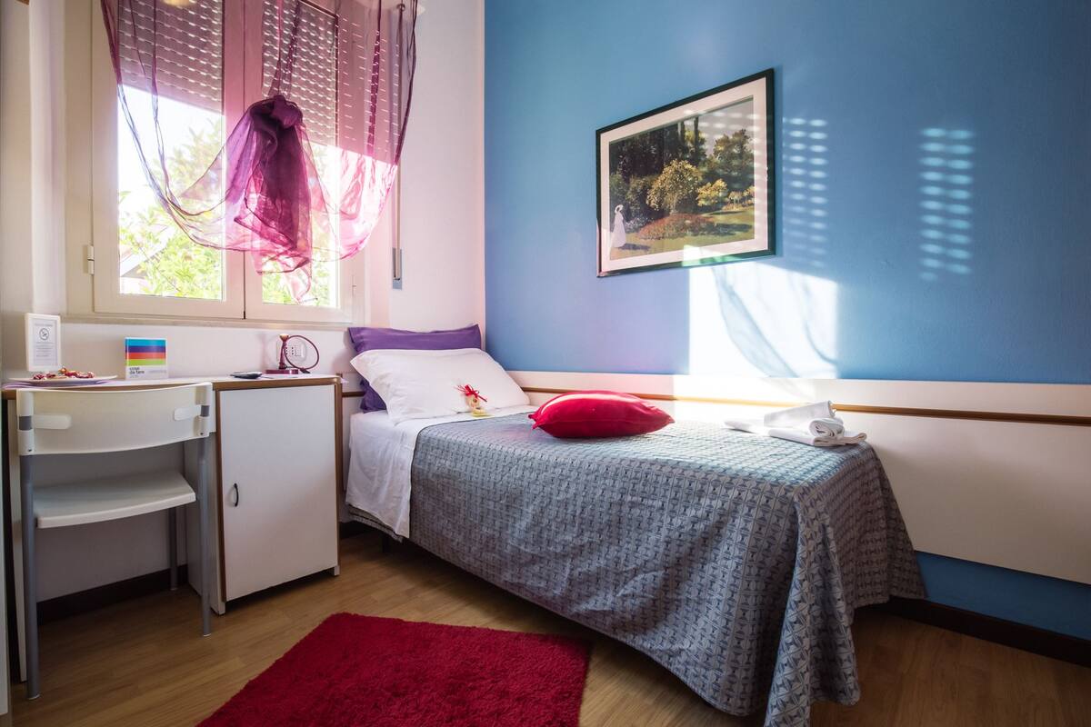 A bedroom with a bed, a desk, a chair, a red rug, a window with sheer curtains, and a framed picture on the wall.