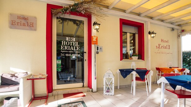 A hotel entrance with a red-framed door, a tiled patio area, and outdoor seating with tables and chairs. - Hotel Eriale (Rimini)