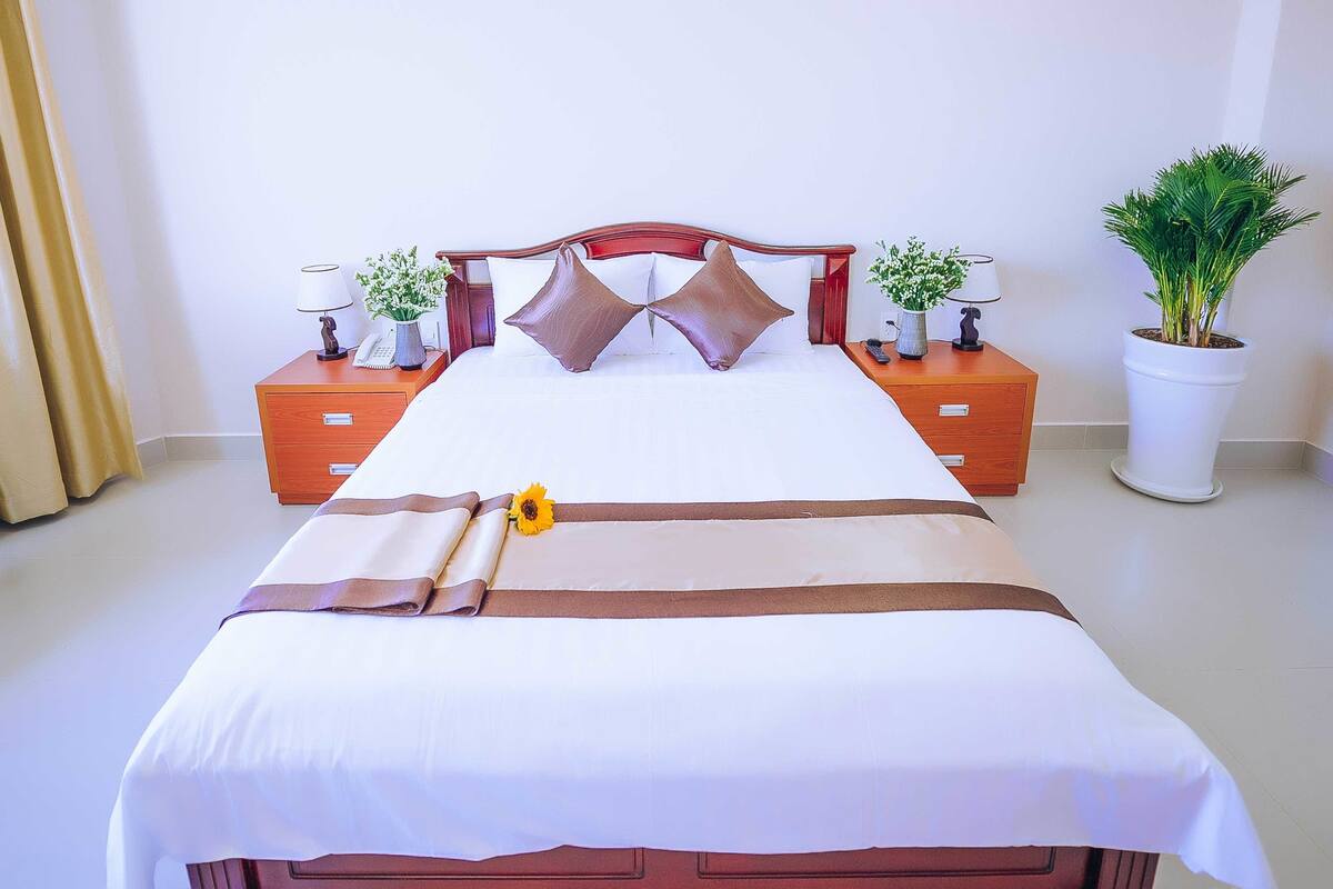 A neatly made bed with a white and brown bedspread, two decorative pillows, a small potted plant, and a nightstand with a lamp and a phone.