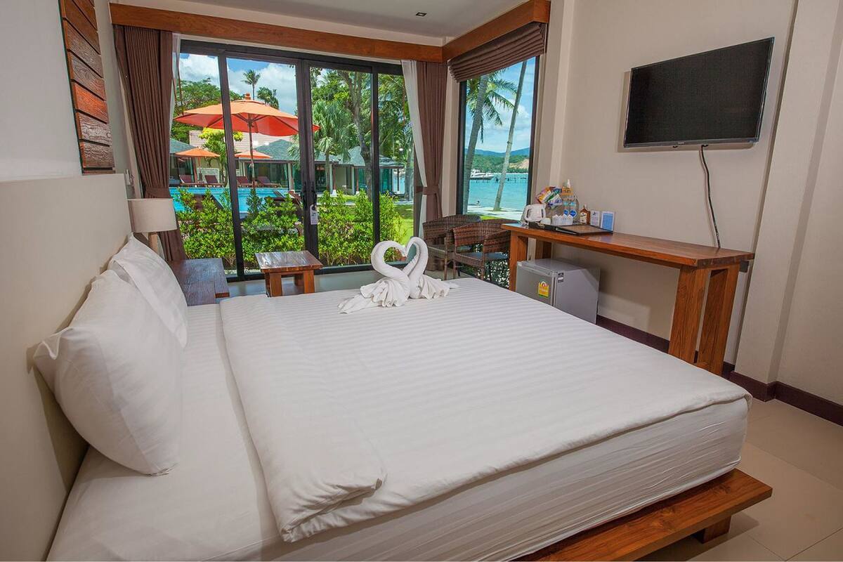 A hotel room with a large bed, a desk with a chair, a TV, and a view of a pool and palm trees.
