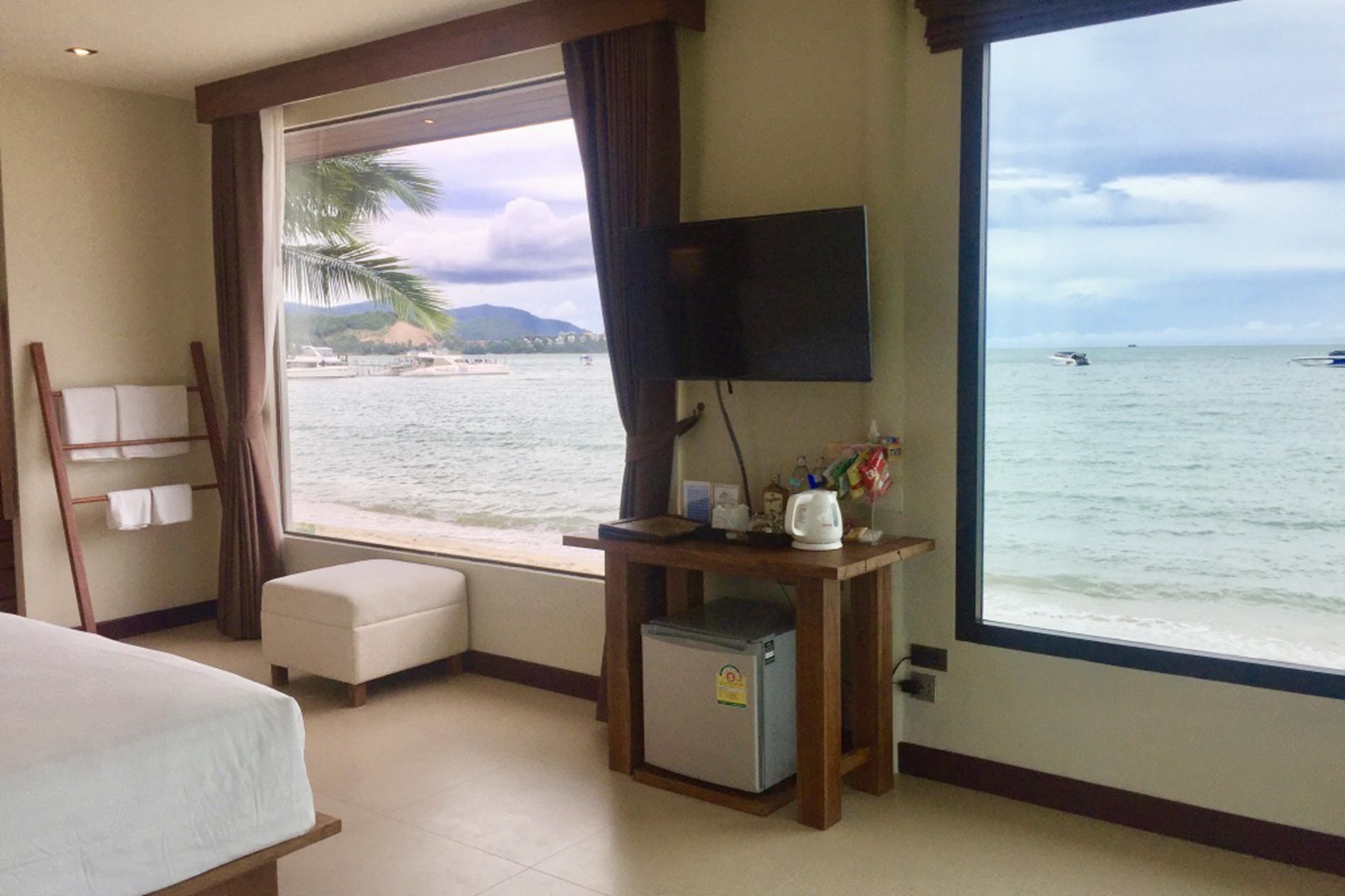 Una habitación de hotel con una cama, una mesita con televisor, un refrigerador y un ventanal con vista al mar y barcos.