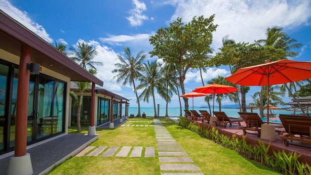 Een resort met een pad naar een zwembad, omgeven door palmbomen en ligbedden met rode parasols.