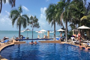 A pool area with lounge chairs and umbrellas, surrounded by palm trees and a view of the sea.
