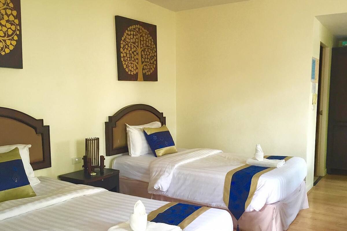 A hotel room with two beds, each with a wooden headboard, white bedding, and blue and gold accents. There is a nightstand with a lamp and a wall-mounted clock.