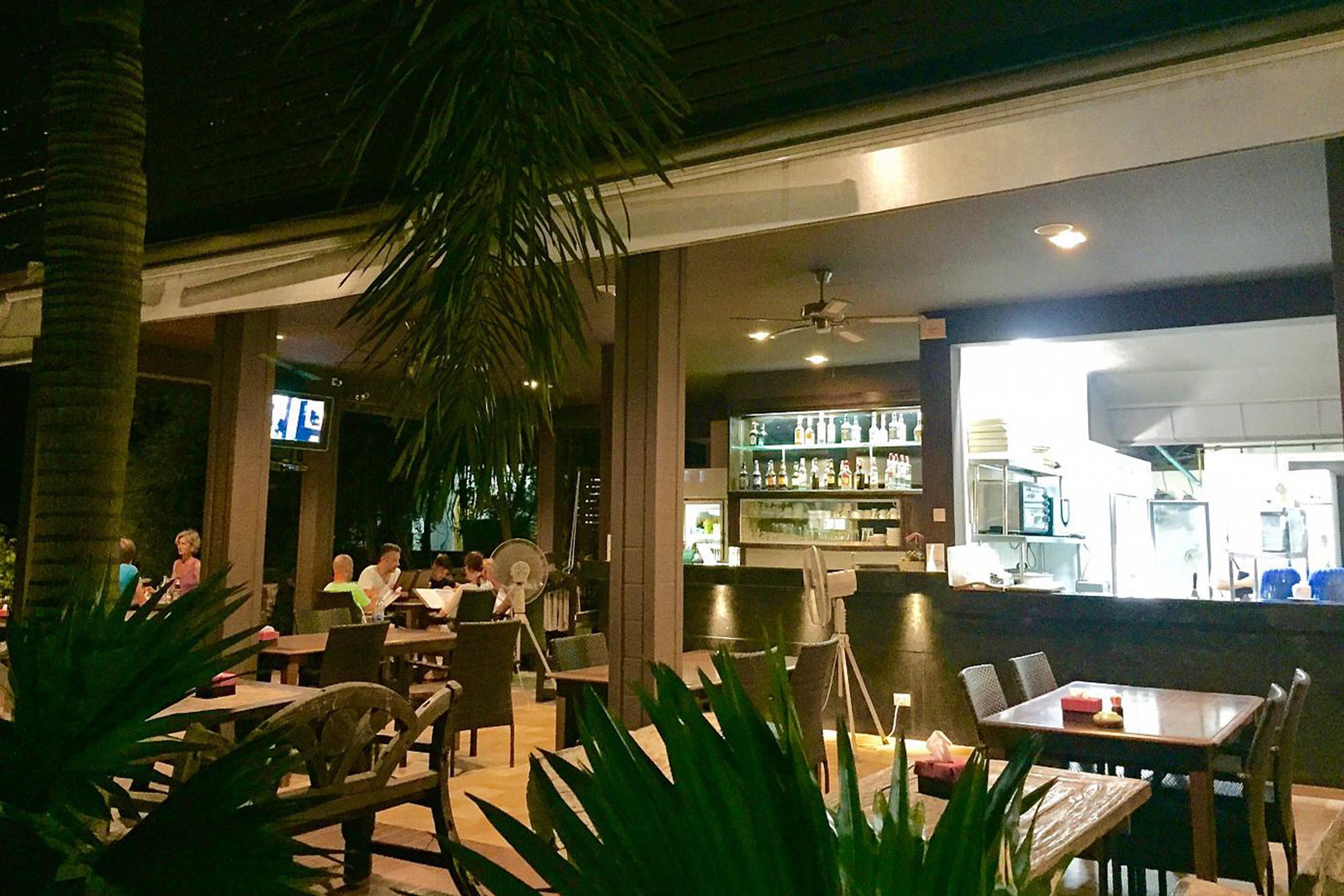 Un espace extérieur aménagé pour les repas, avec des tables et des chaises, un bar doté d’un comptoir et d’étagères remplies de bouteilles, et une cuisine visible au fond.