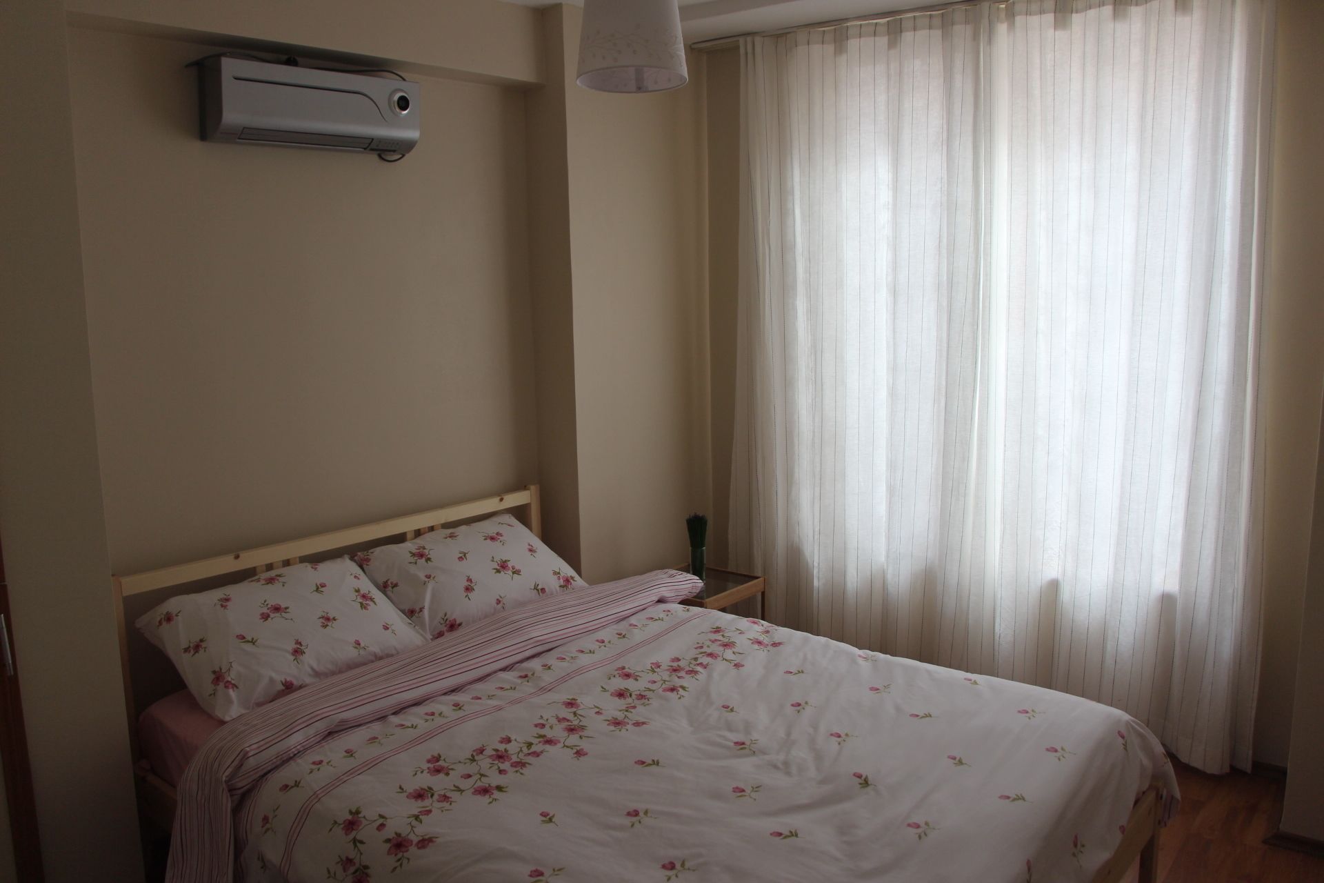 Un dormitorio con cama, mesita de noche, aire acondicionado de pared y ventana con cortinas.