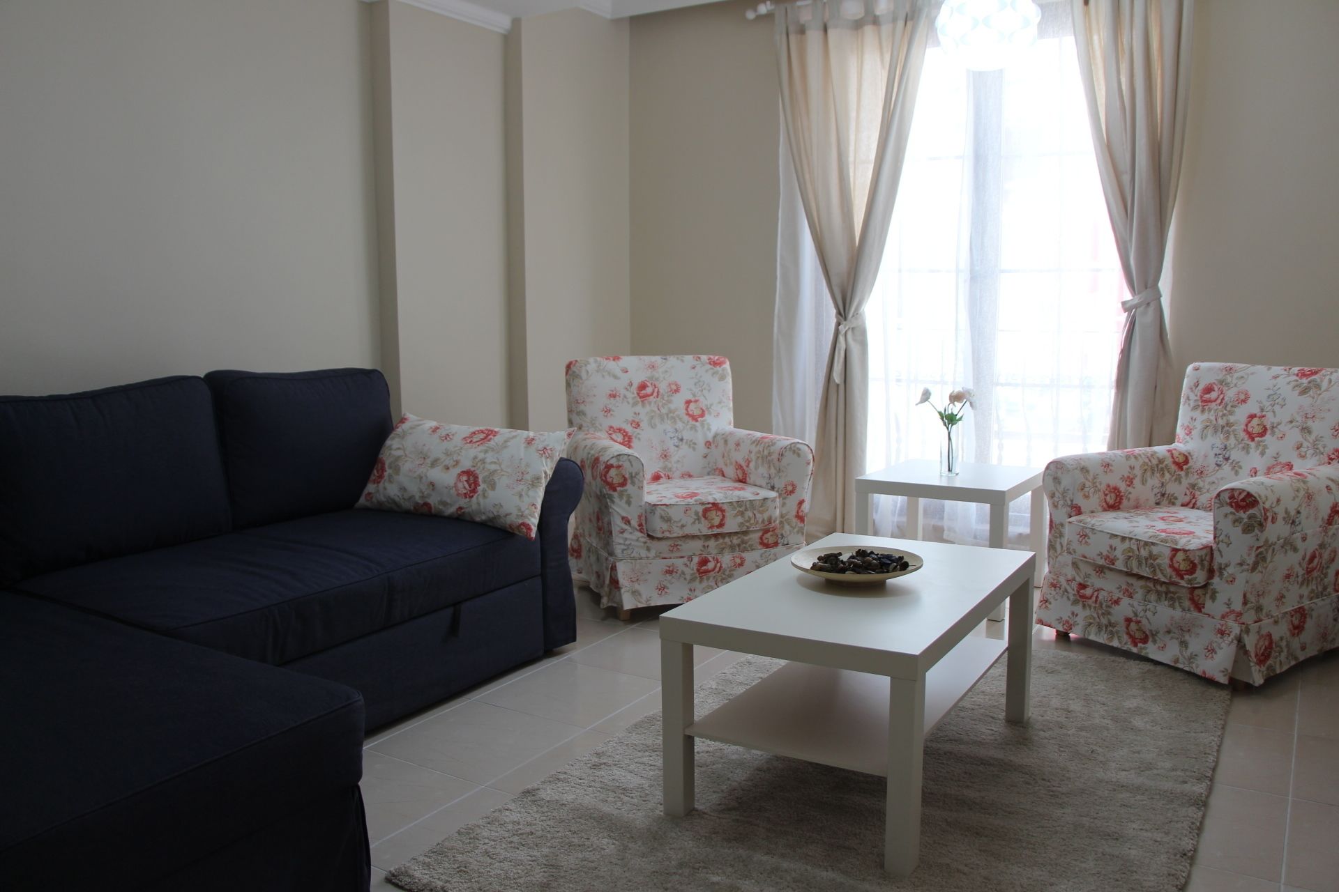 A living room with a dark sofa, two floral armchairs, a white coffee table, and a small table with a vase of flowers.