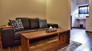 A modern living room with a dark leather sofa, patterned cushions, a wooden coffee table, and a view into a kitchen area. - Chika Odoru (Kyoto)