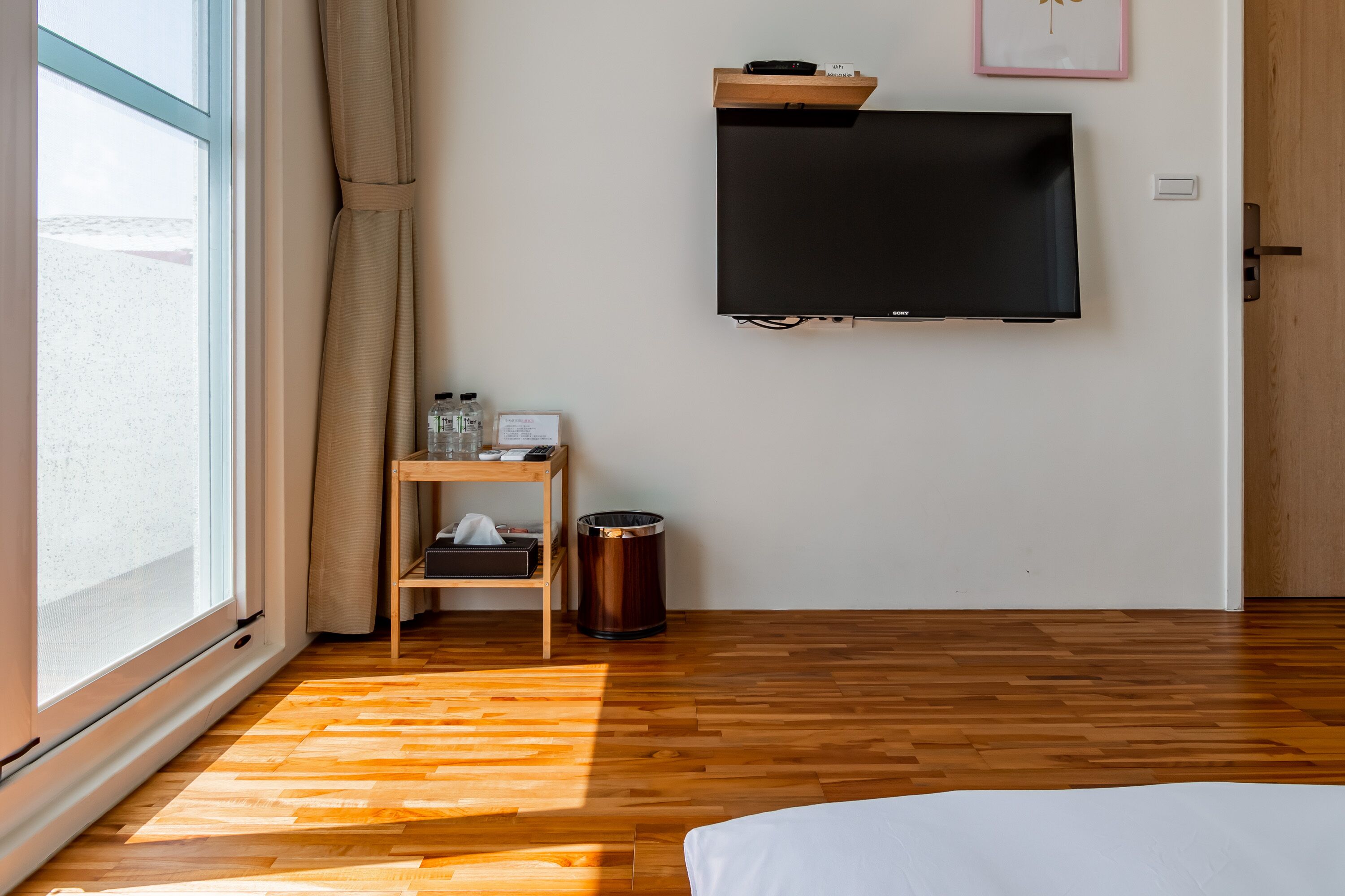 Habitación de hotel con suelo de madera, cama, televisor montado en la pared, una mesita con un cesto de basura y una ventana con cortinas.