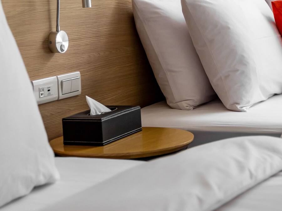 A neatly made bed with white bedding, a bedside table with a tissue box, a wall-mounted night light, and a power outlet.