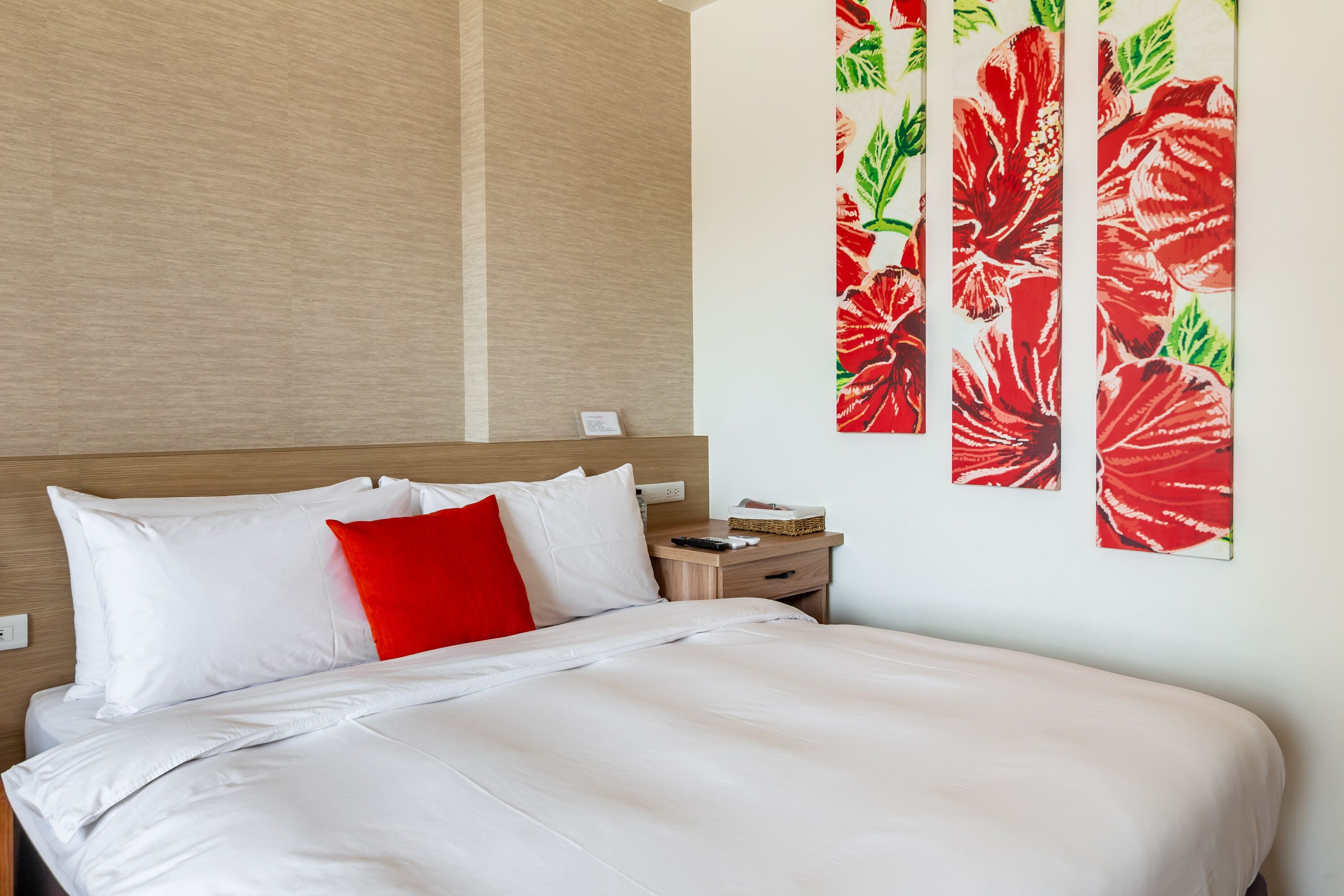 A neatly made bed with white and red bedding, a wooden headboard, and a nightstand with a remote control and a basket.