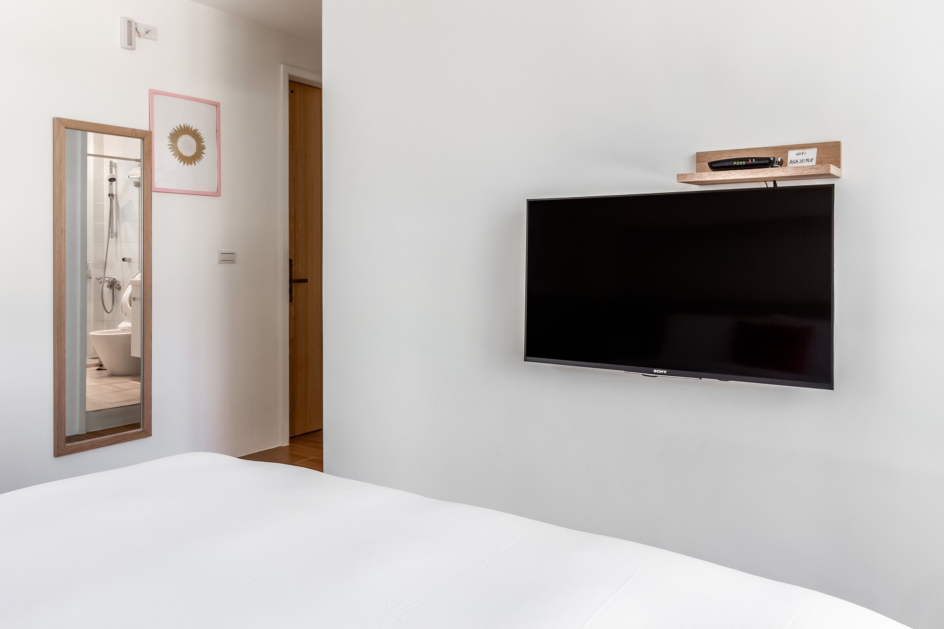 Un dormitorio con una televisión de pantalla grande montada en la pared, un estante de madera con un control remoto y un espejo que refleja un baño con bañera.
