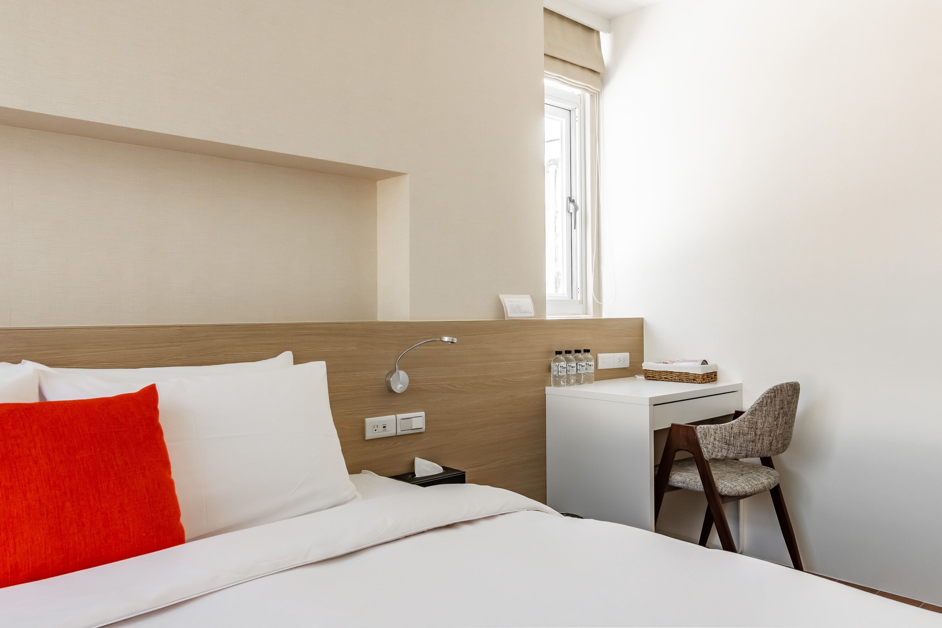A hotel room with a bed, a desk, a chair, a window, and a bedside table with a lamp and bottles.