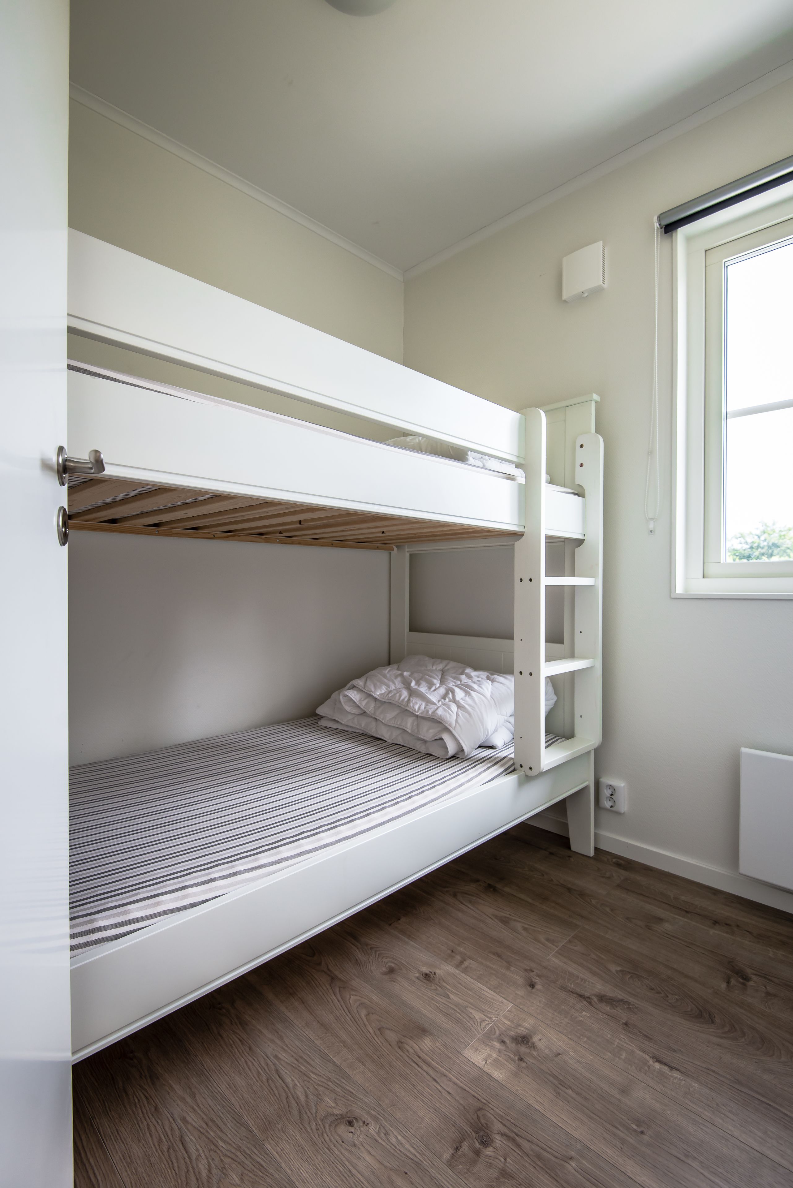 A bunk bed with a wooden headboard, a white wall, and a window with blinds.