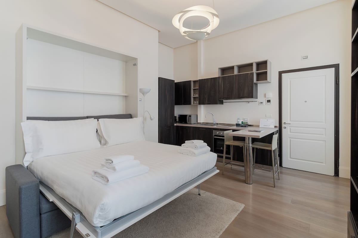 A modern bedroom with a bed, bedside tables, a desk, and a kitchenette.