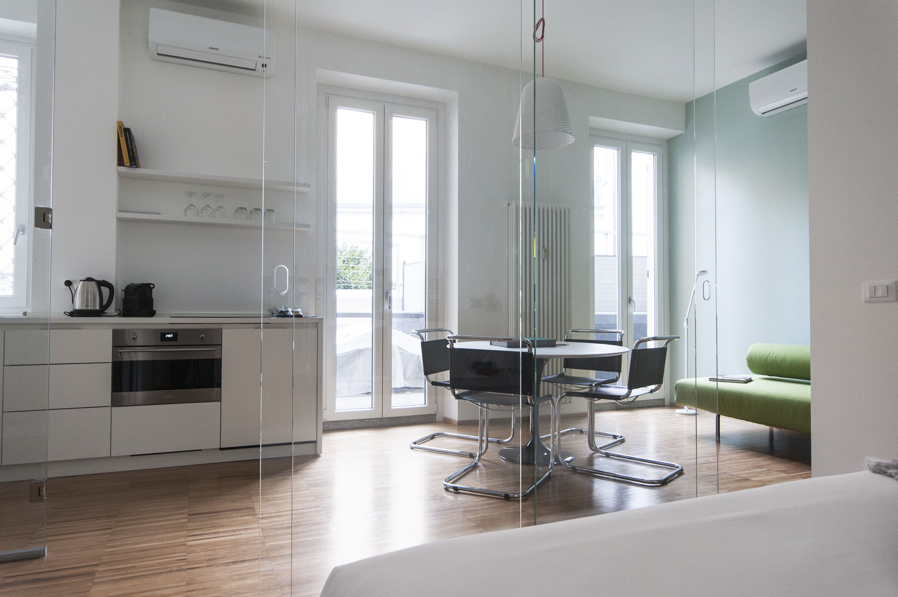 Una cocina moderna con horno empotrado, una zona de comedor con mesa y sillas de vidrio, y un sofá verde en una habitación luminosa con ventanas amplias.