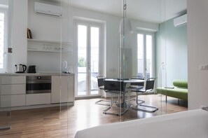 Eine moderne Küche mit eingebautem Ofen, eine Essecke mit Glastisch und Stühlen sowie ein grünes Sofa in einem hellen Raum mit großen Fenstern.