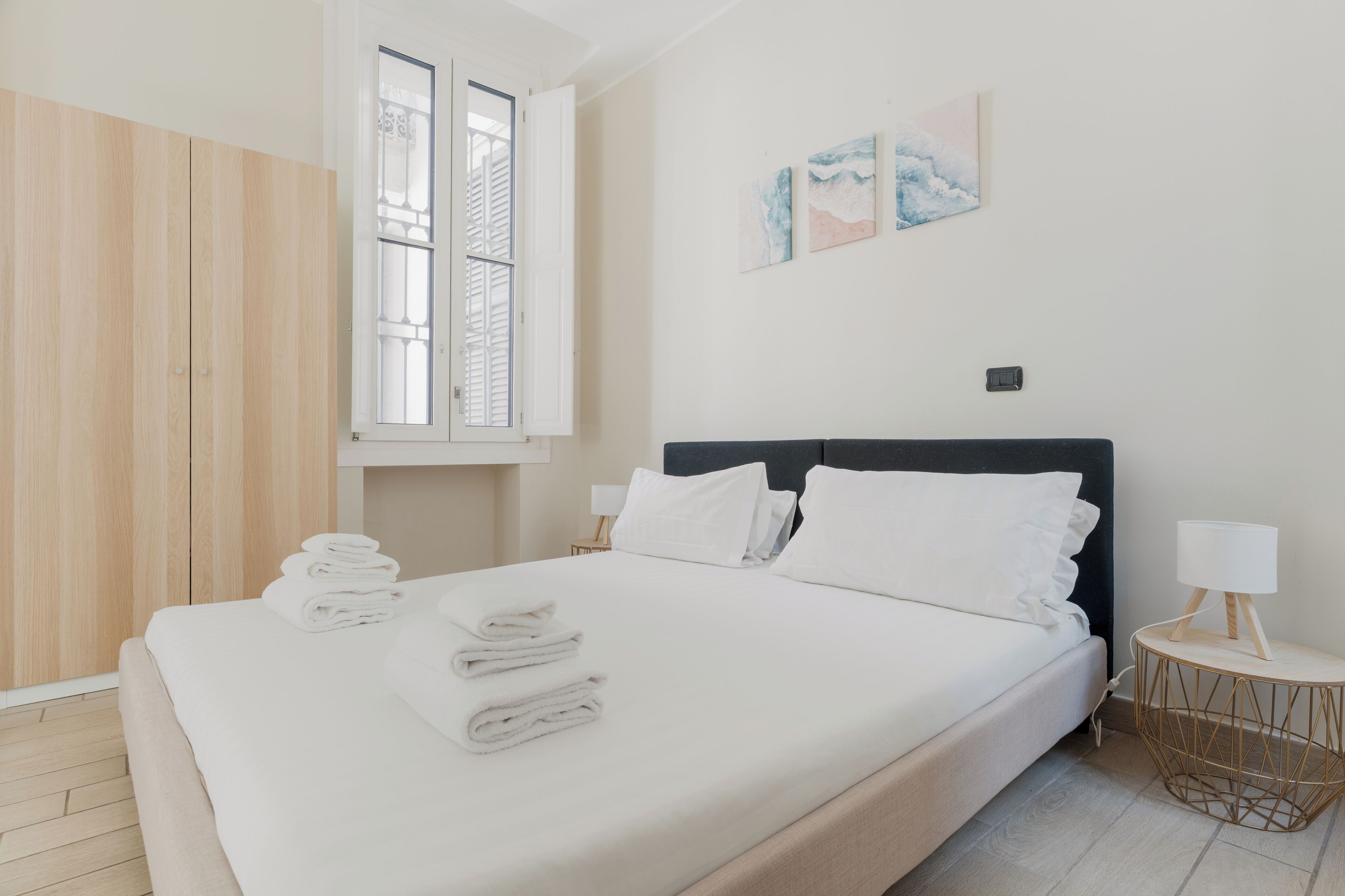 Ein ordentlich bezogenes Bett mit weißen Leinentüchern, ein Fenster mit Rollläden, ein an der Wand hängendes Kunstwerk und ein Nachttisch mit Lampe.