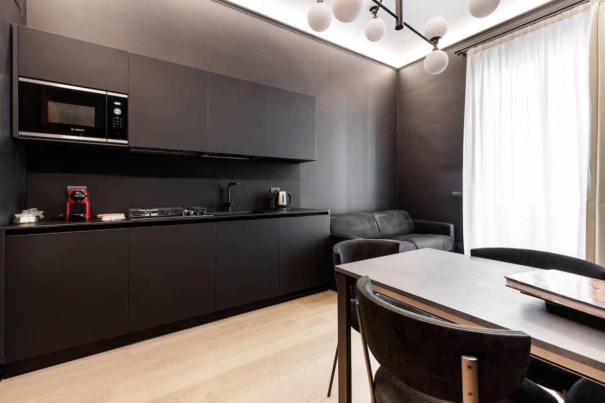 A modern kitchen with dark cabinetry, a microwave, and a coffee machine. There is a dining table with chairs and a sofa in the adjacent living area.