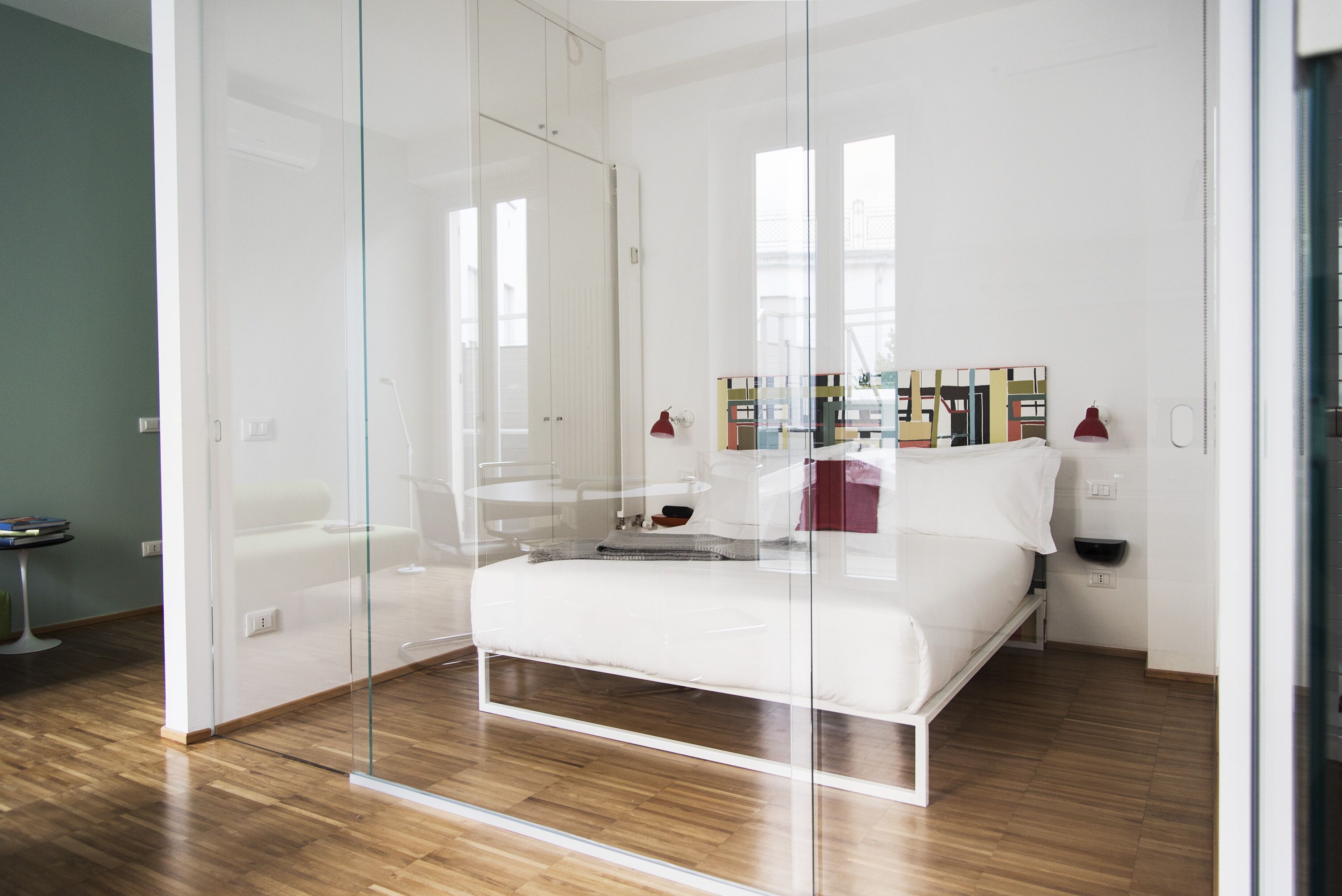 A modern bedroom with a glass partition, a wooden floor, a bed, a bedside table, and a small seating area.
