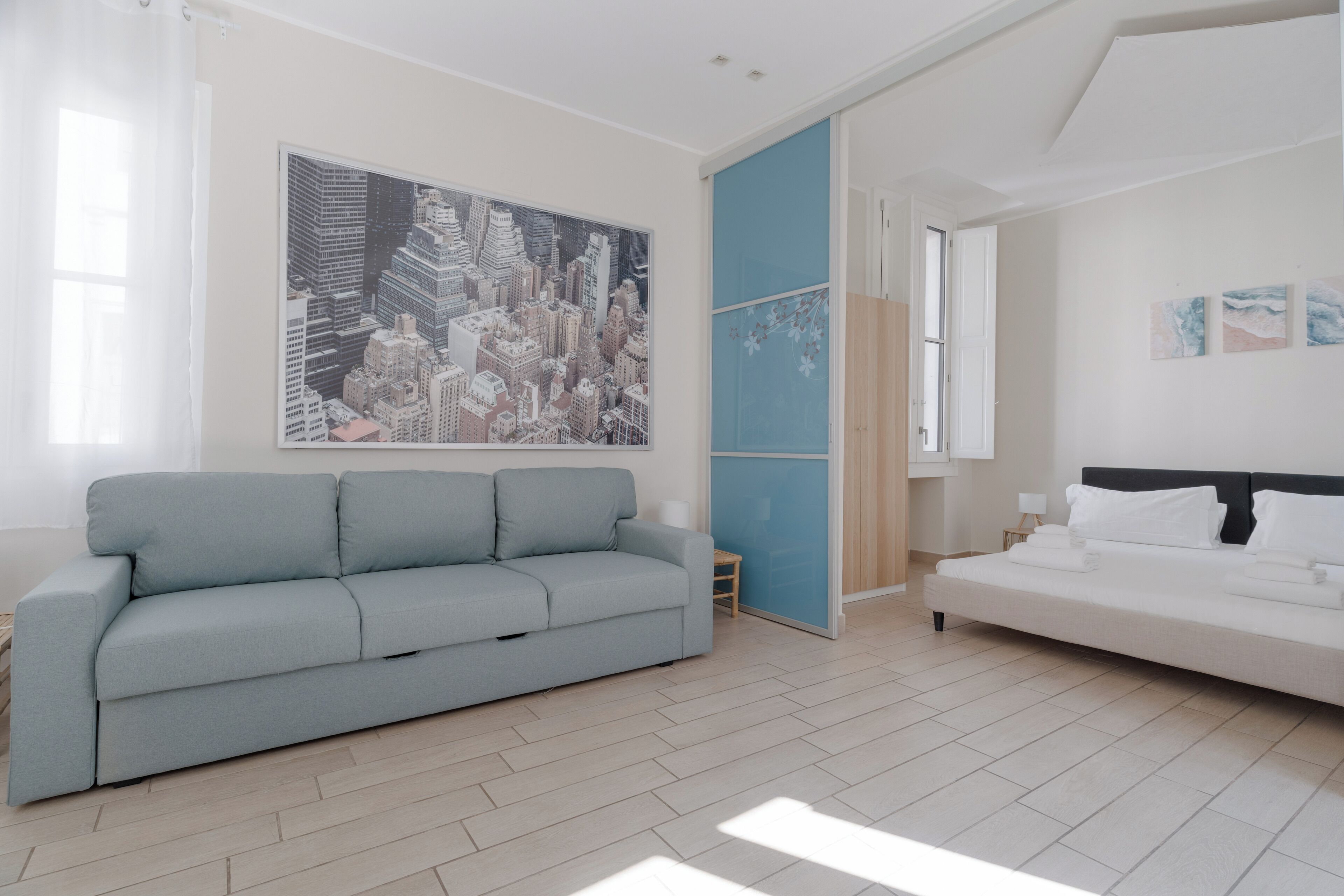 Una habitación moderna con un sofá azul claro, una cama blanca, un cuadro grande de un paisaje urbano y un separador azul.