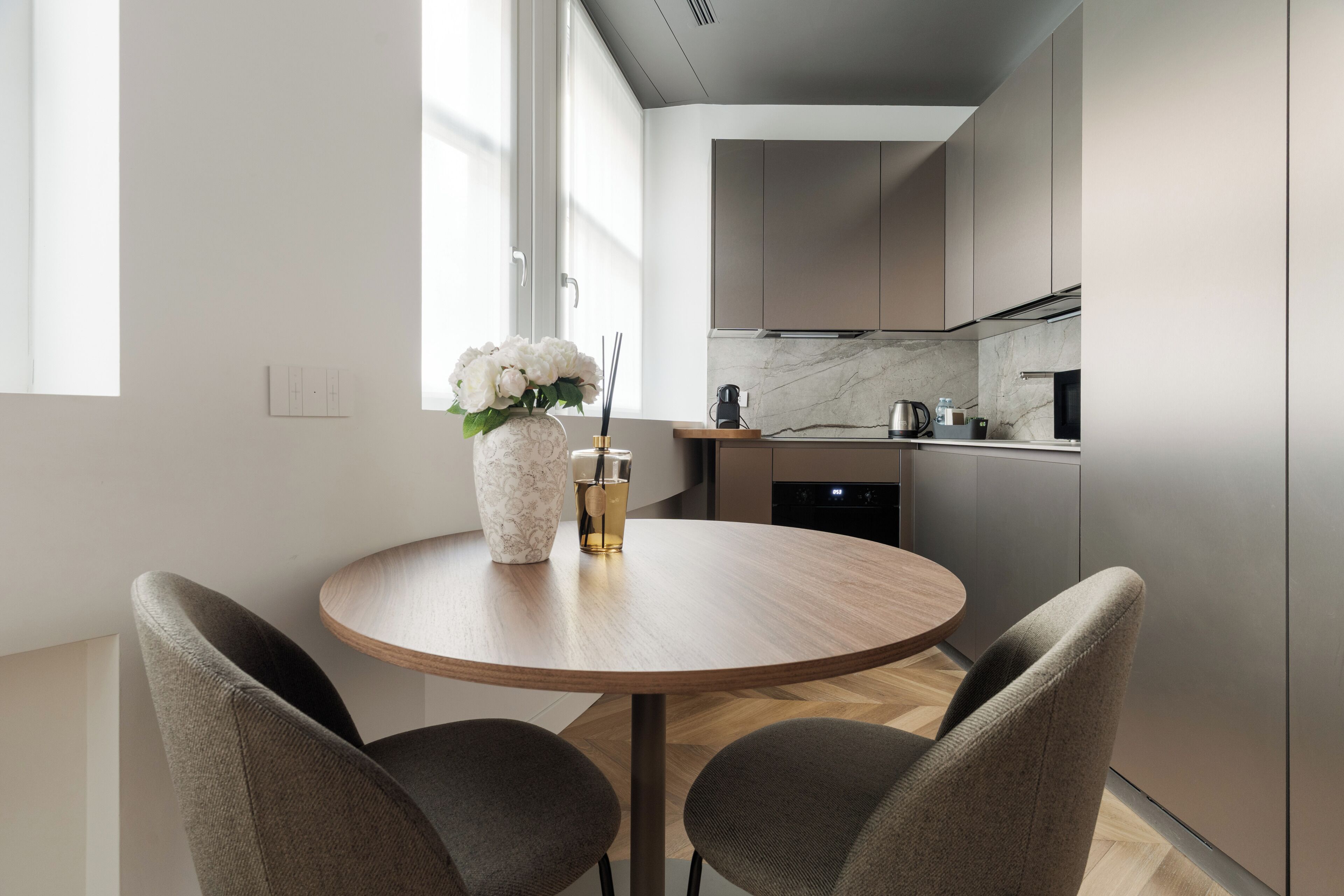 A modern kitchen with a round dining table, two chairs, a vase with flowers, and a coffee maker.