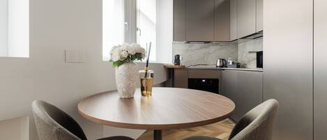 A modern kitchen with a round dining table, two chairs, a vase with flowers, and a coffee maker.