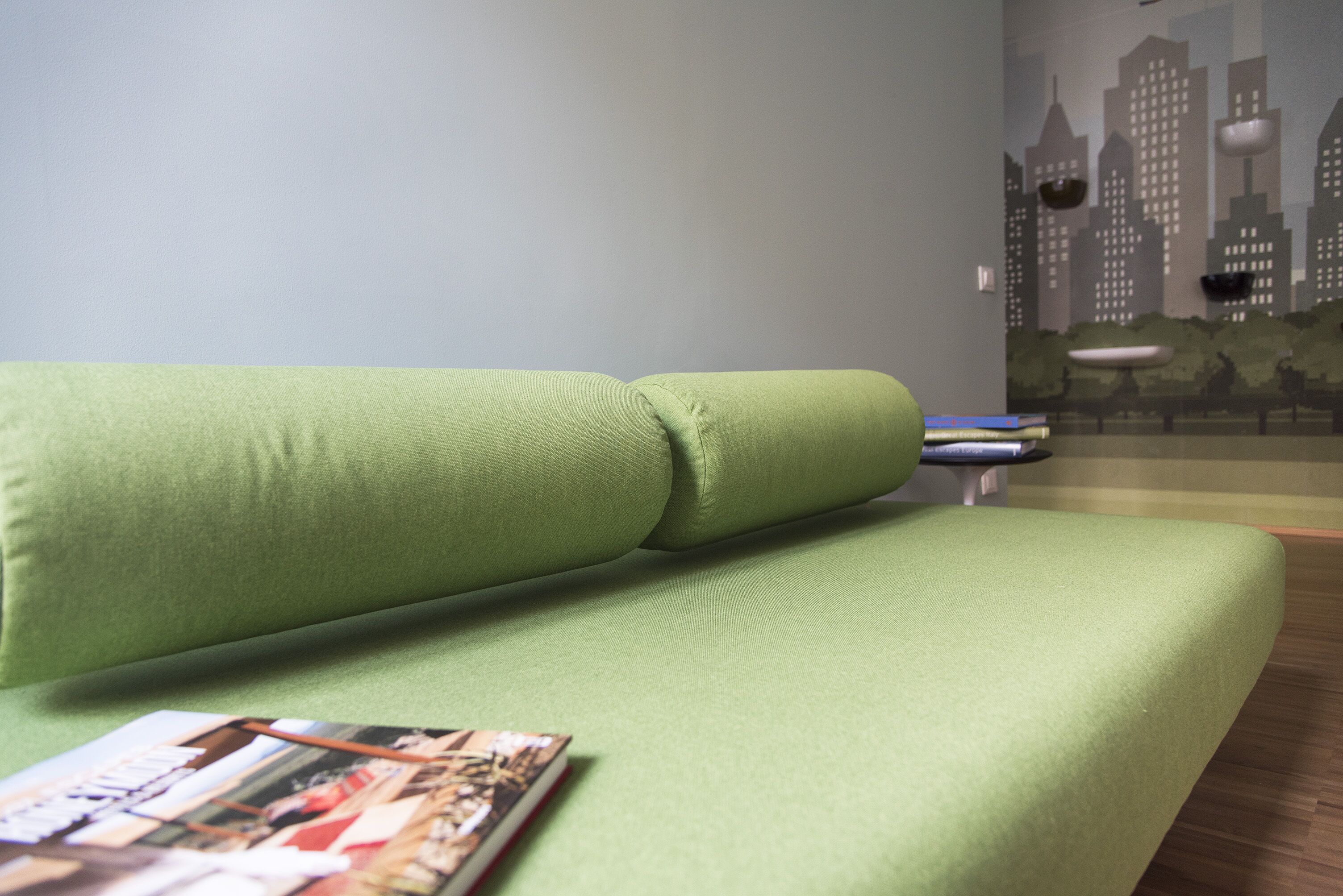 Un canapé vert avec un magazine dessus, une fresque représentant un paysage urbain sur le mur et un plancher en bois.