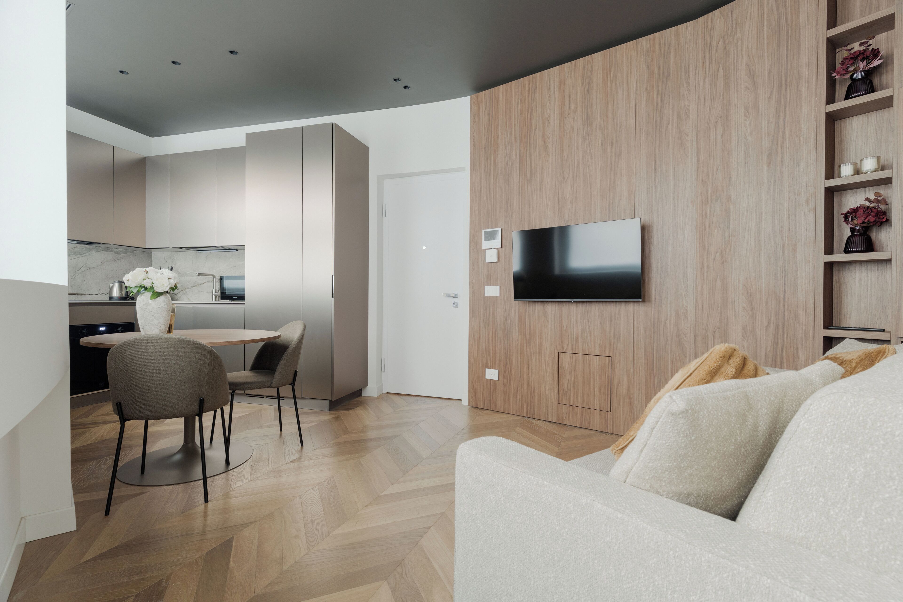A modern living room with a sofa, a flat-screen TV mounted on a wooden wall, a dining area with a table and chairs, and a kitchen in the background.