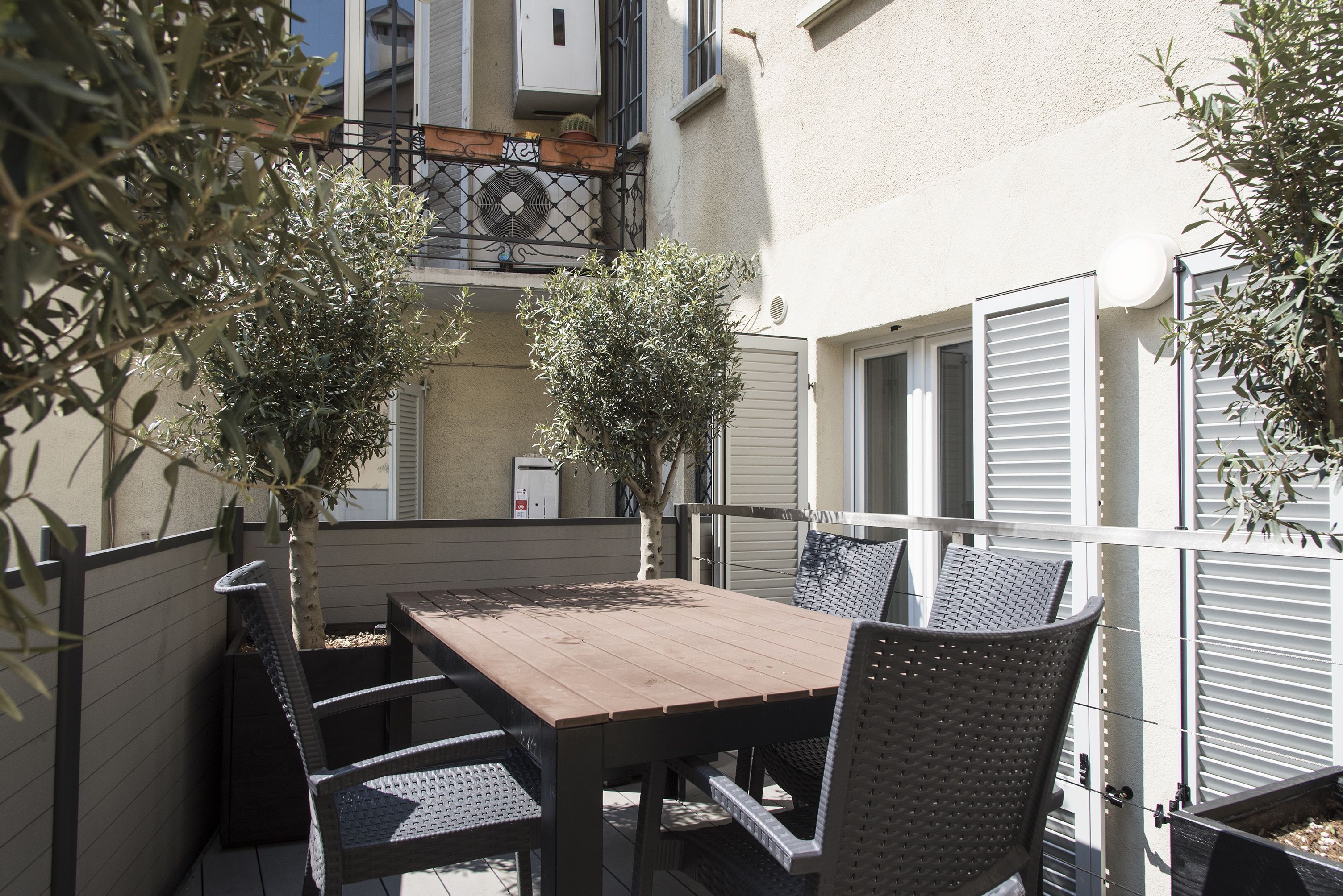 Een terras buiten met een houten tafel en rieten stoelen, omringd door olijfbomen in potten.