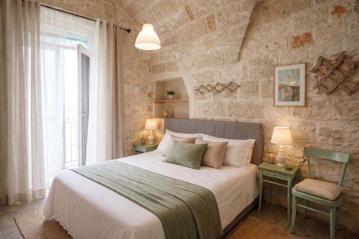 A bedroom with a stone wall, a bed with a green blanket, two bedside tables with lamps, and a window with curtains.