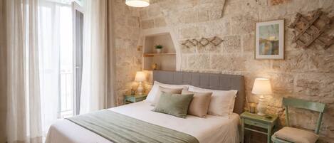 A bedroom with a stone wall, a bed with a green blanket, two bedside tables with lamps, and a window with curtains.
