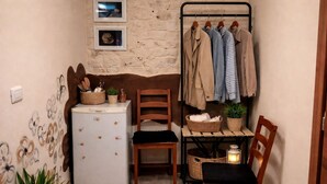 Une pièce de taille modeste, avec un réfrigérateur blanc, deux chaises en bois, une petite table avec des paniers, un porte-manteau fixé au mur et une plante en pot.