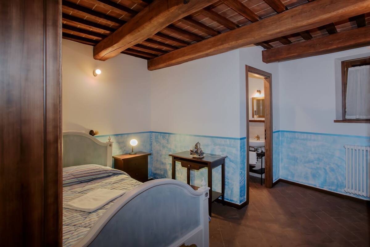 Een slaapkamer met een bed, nachtkastje en een wandlampje. Er is een houten plafond en een raam met gordijn.