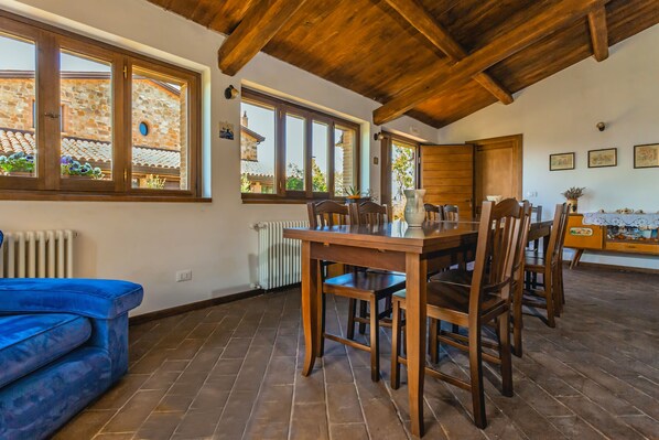 A dining area with a wooden table and chairs, large windows, and a blue sofa. - Agriturismo Eutopia (San Leo)