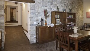 A rustic interior with a stone wall, wooden ceiling beams, a dining area with a table and chairs, and a bookshelf. - Agriturismo Eutopia (San Leo)