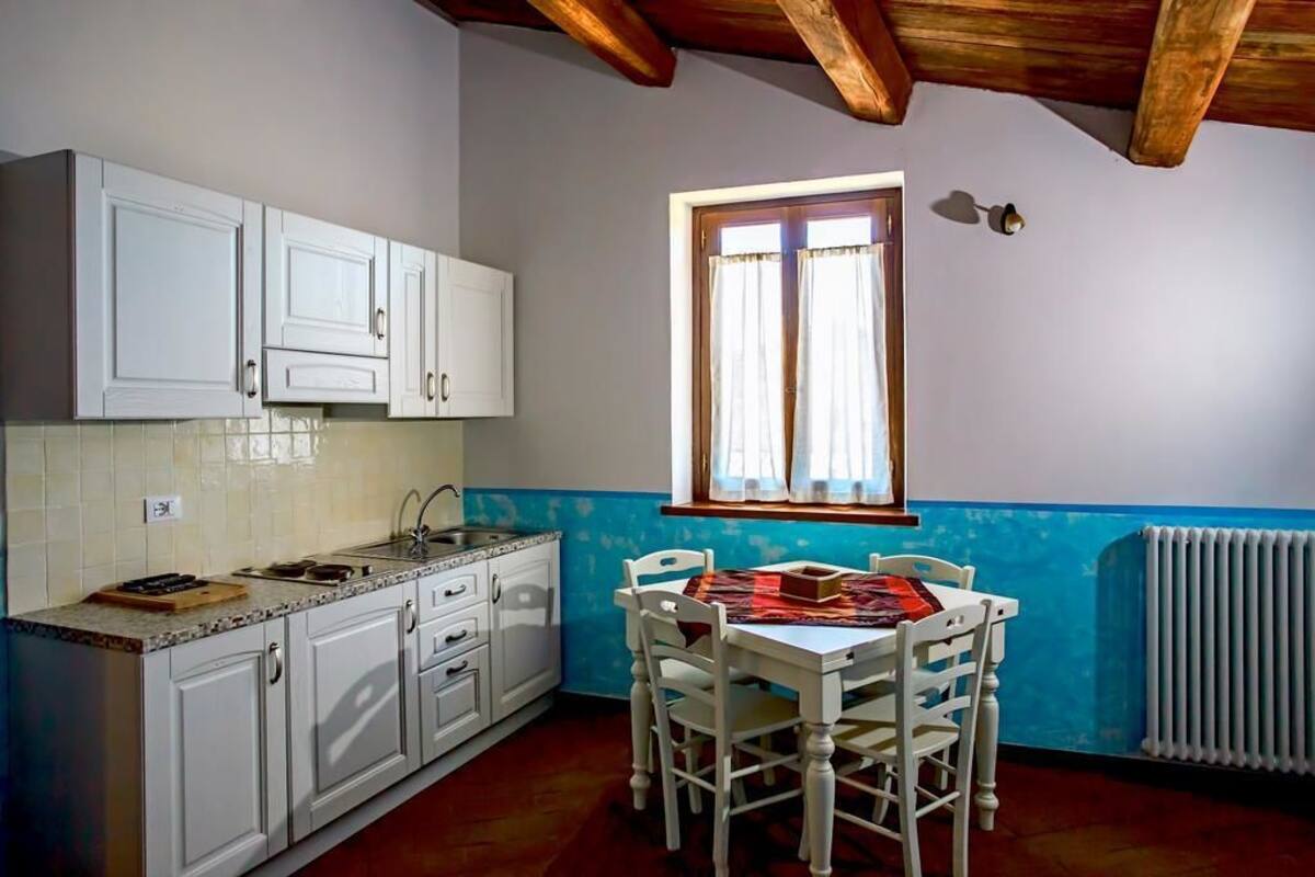 Een keuken met witte kastjes, een raam met gordijnen en een tafel met stoelen.