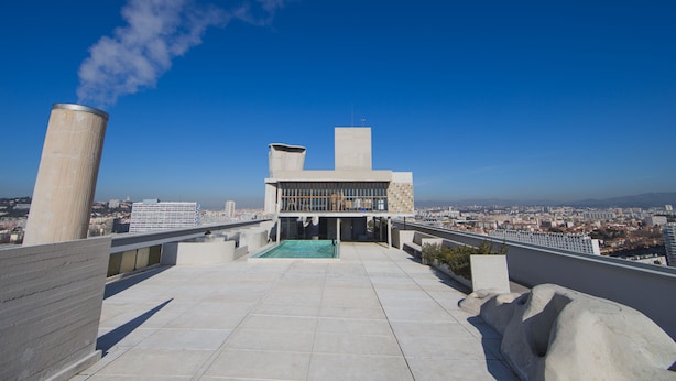 A rooftop terrace with a view of the city, a large cylindrical structure, and a building with a flat roof. - Hôtel Le Corbusier (Marseille)