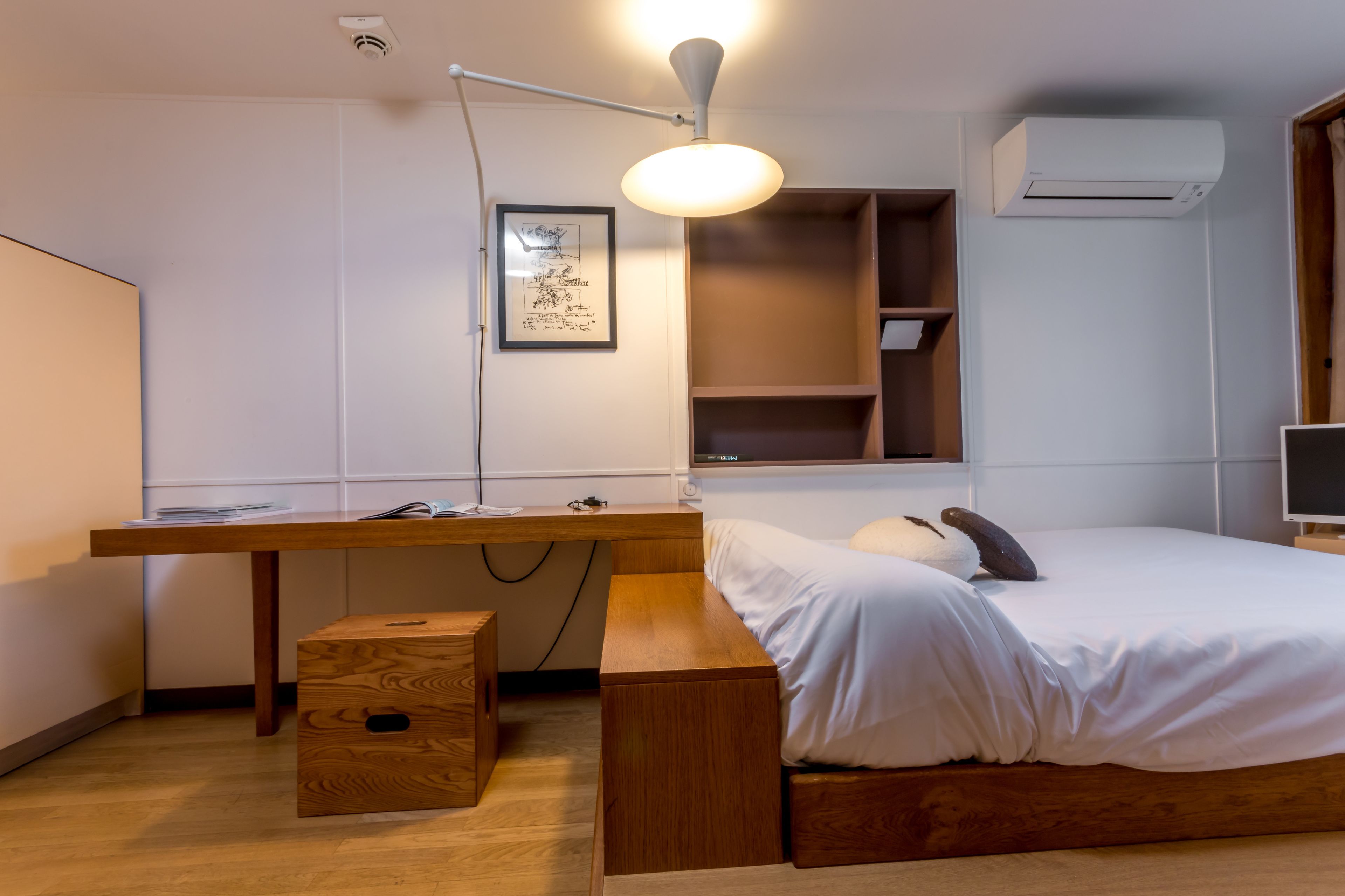 A bedroom with a wooden desk, a bed, a bedside table, a hanging light, and a wall-mounted air conditioner.