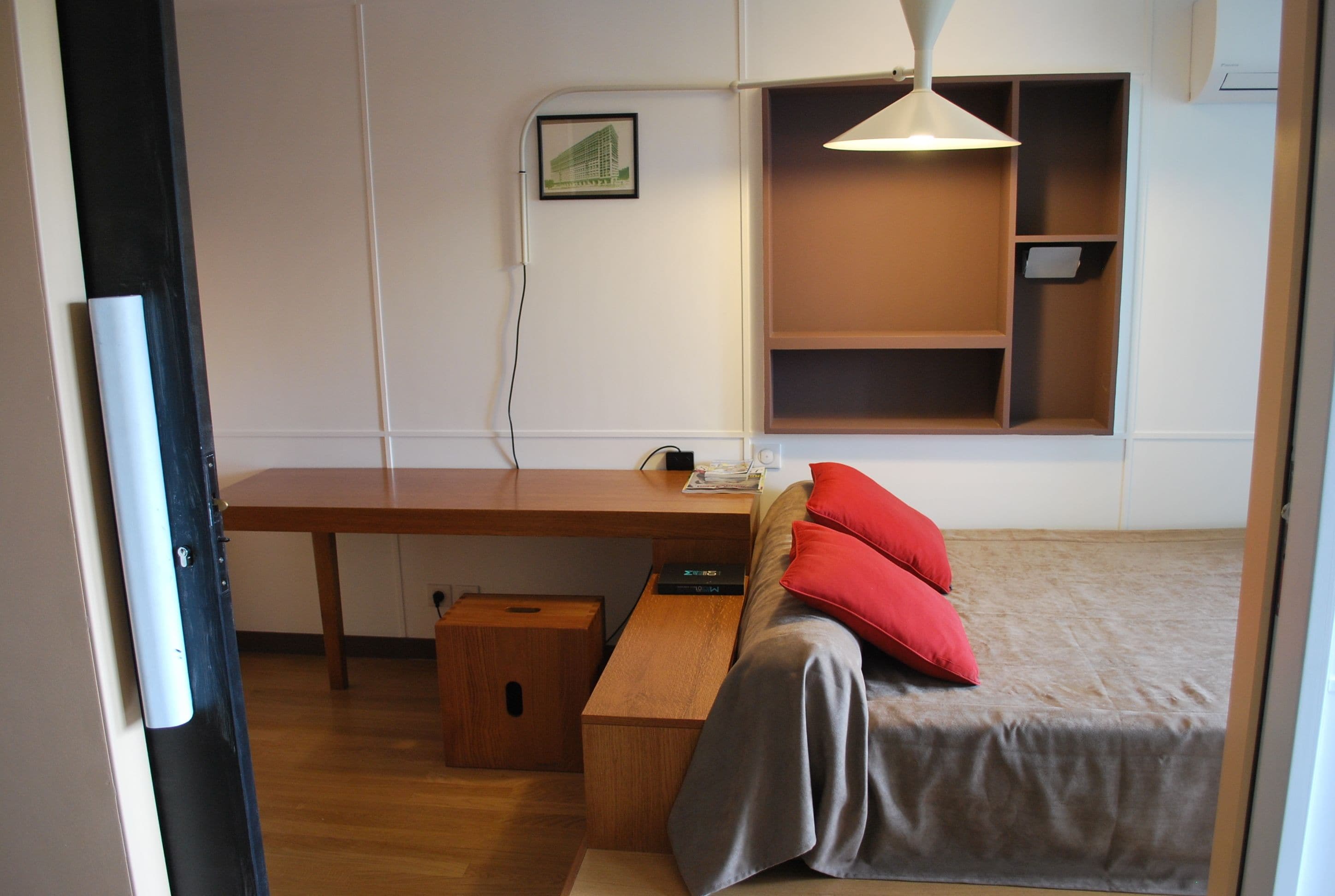 A compact room with a wooden desk, a bed with red pillows, a small wooden stool, and a wall-mounted shelf.