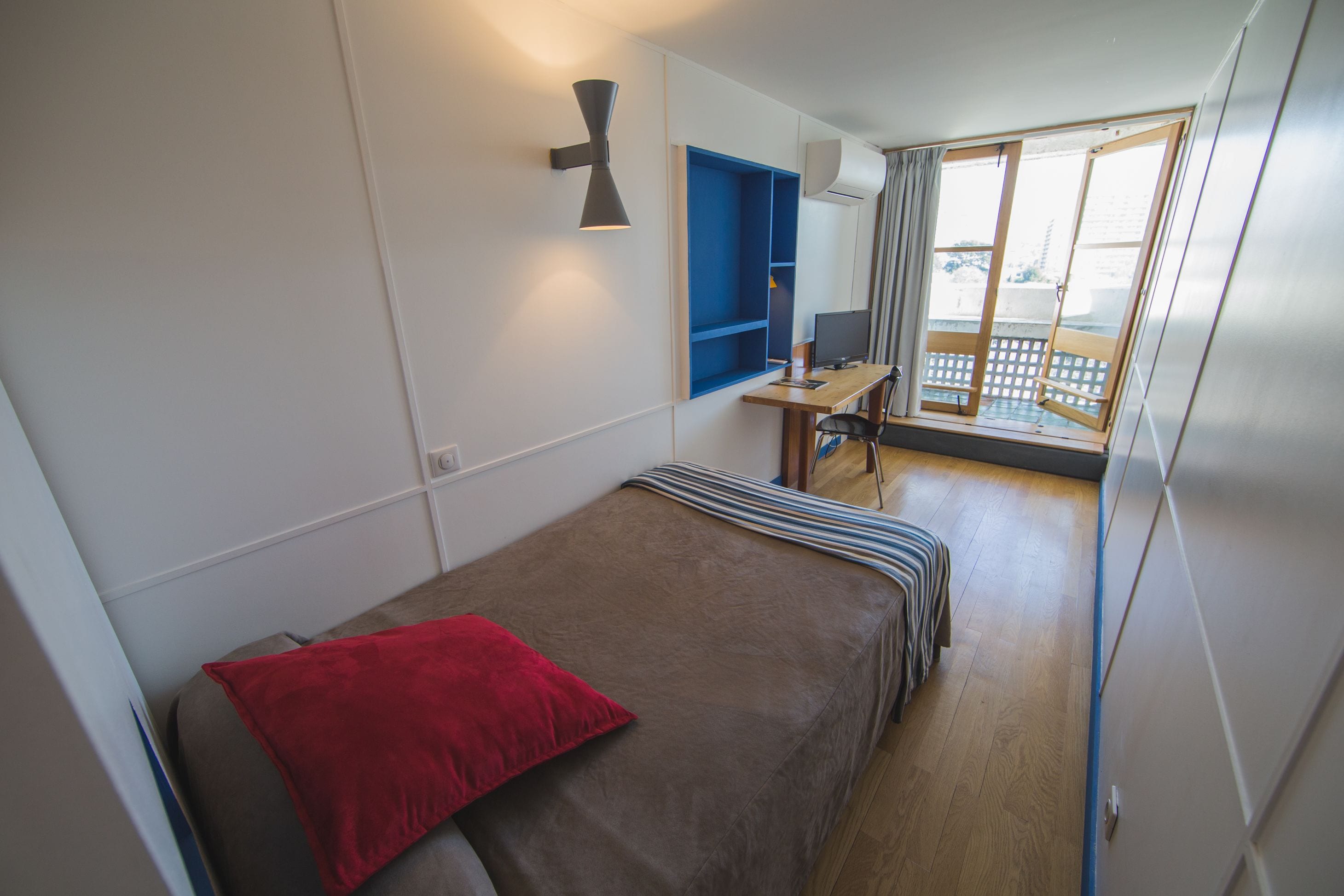 A hotel room with a bed, a desk with a computer, a window with a view, and a wall-mounted air conditioner.