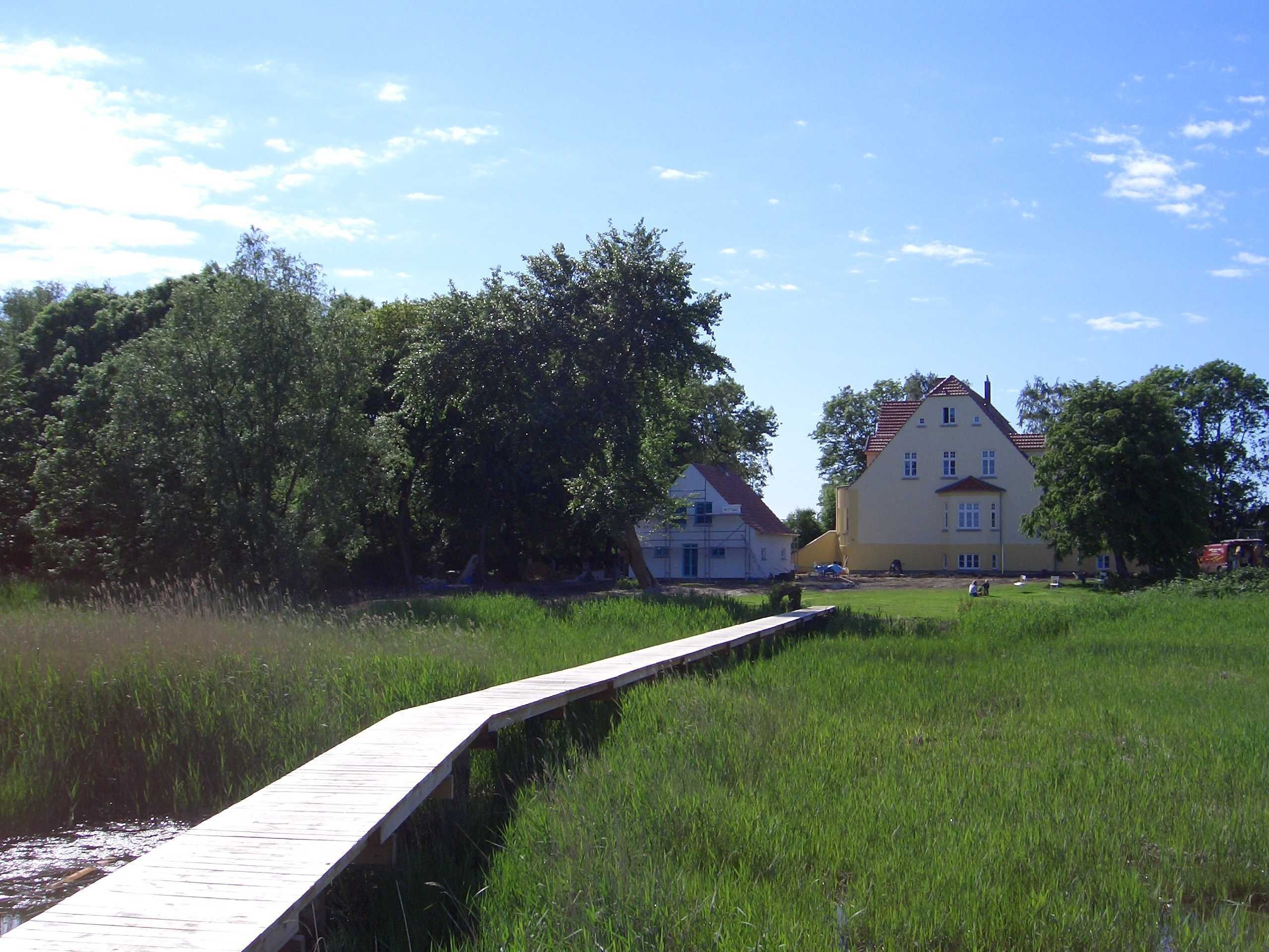 A wooden bridge over a grassy field leading to a large yellow building with a red roof, surrounded by trees.
