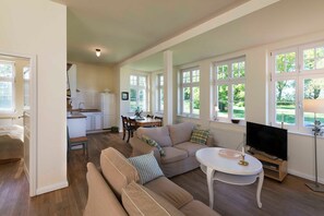 Un salon spacieux avec un canapé beige, une table à manger et un téléviseur. Au fond, on aperçoit une cuisine avec des meubles blancs et une table à manger.