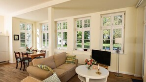Una sala de estar con sofá, una mesa de comedor con sillas, televisor y ventanas amplias con vistas a la vegetación.