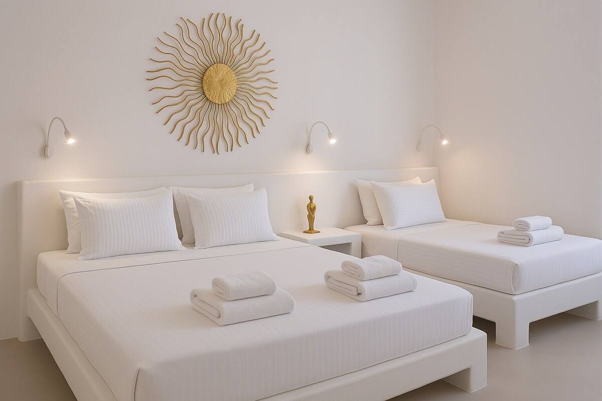 Quarto de hotel com duas camas, roupa de cama branca, uma arte decorativa na parede que lembra um sol e luminárias de cabeceira.